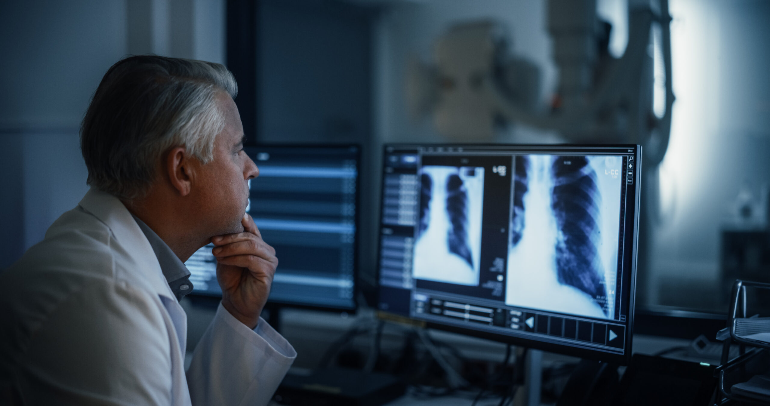 Experienced Radiographer Examines Chest X-rays on Multiple Computer Monitors in a Dark Medical Room. Modern Diagnostic Equipment Ready to Take Medical Imaging Scans of a Male Patient