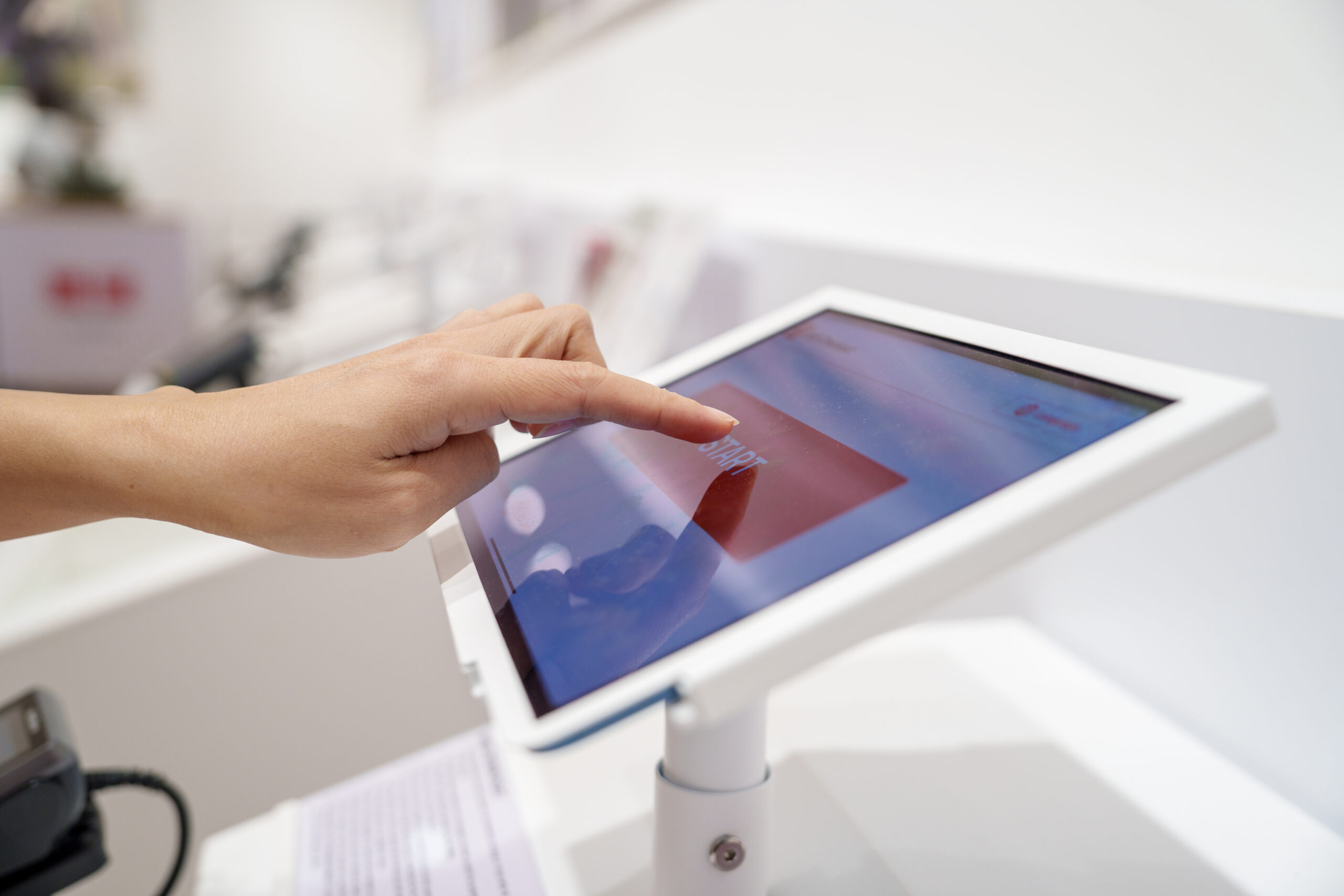 A close-up view of a person’s hand interacting with a touchscreen kiosk in a retail environment, showing the action of tapping the start button on the display.