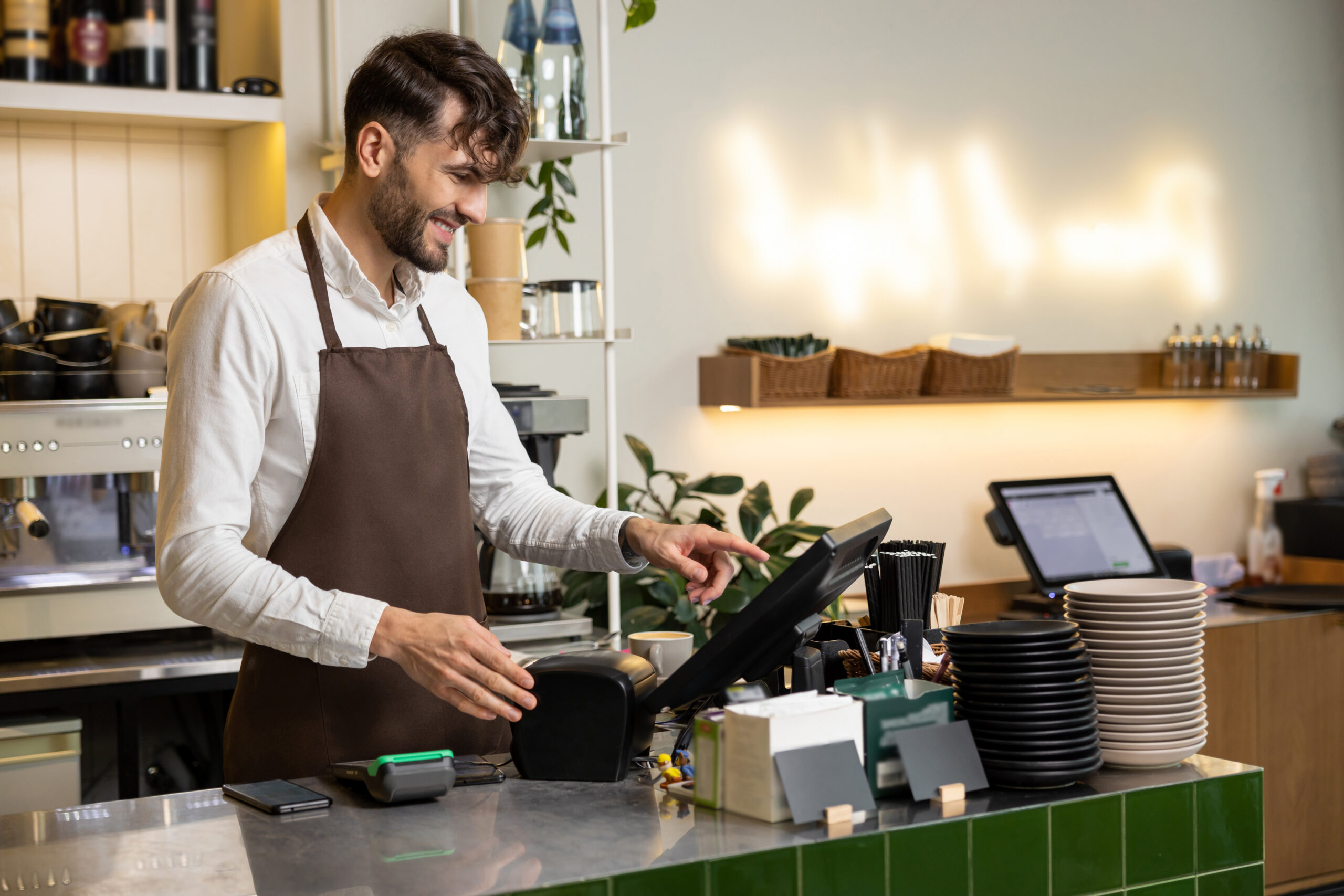 Man waiter in apron working in coffee shop using terminal while standing at counter